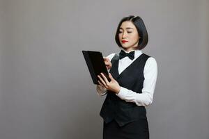 Jeune asiatique serveuse dans uniforme vérification café menu sur numérique tablette. restauration un service Personnel gérant en ligne commande et tapoter sur écran tactile tandis que posant dans studio sur gris Contexte photo