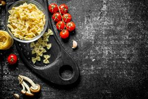 brut farfalle et tagliatelles pâte dans une bol sur une Coupe planche avec champignons, tomates et épices. photo