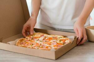 à emporter aliments. une femme détient une papier carton boîte avec prêt à manger Pizza sur le table dans le cuisine. photo