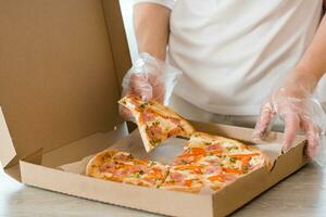 à emporter aliments. une femme dans jetable gants prend une tranche de Pizza de une papier carton boîte sur le table dans le cuisine. photo