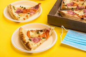 à emporter et livraison. une tranche de Pizza dans une jetable Plastique plaque, une boîte de Pizza et une protecteur masque sur une Jaune Contexte photo