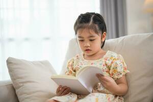 joyeux content asiatique enfant bébé fille souriant et en train de lire livre tandis que séance sur canapé canapé dans vivant pièce à maison. fille relex en train de lire livre sourire à canapé dans le maison. retour à école concept. photo