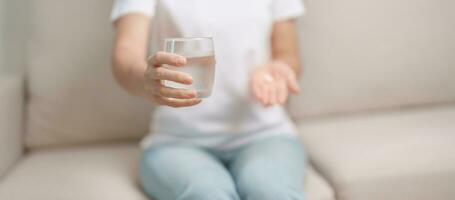 femme main en portant médicament anti douleur pilule et l'eau verre sur le canapé à maison, prise pour maux de tête, estomac mal, la diarrhée douleur de nourriture empoisonnement, l'endométriose, hystérectomie et menstruel photo