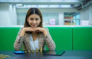 magnifique asiatique affaires femme est séance avec sa menton sur sa main à la recherche à le caméra avec sourire tandis que sa bureau a une tablette, calculatrice et piles de or pièces de monnaie. photo