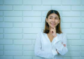 magnifique asiatique affaires femme est permanent dans une réfléchi pose avec une sourire à le copie espace avec une blanc brique arrière-plan, numérique commercialisation. photo