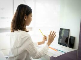 magnifique asiatique en ligne vendeur est en utilisant sa tablette à record produit Commentaires tandis que séance dans sa bureau. photo