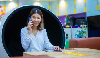 magnifique asiatique affaires femme séance sur moderne rond chaise et en utilisant téléphone à communiquer avec client avec sourire tandis que à la recherche à le caméra, numérique commercialisation. photo