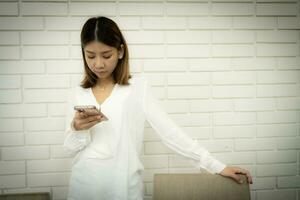 magnifique asiatique Bureau fille est permanent et en portant sa téléphone à vérifier en dehors sa en ligne boutique contre une blanc brique mur Contexte. photo
