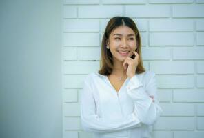 magnifique asiatique affaires femme est permanent contre une blanc brique mur avec sa main sur sa joue et une sourire sur sa affronter, tandis que à la recherche vers le droite côté de le image, numérique commercialisation. photo