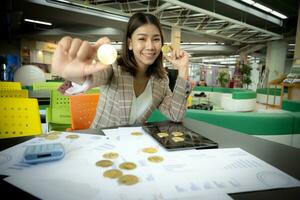 magnifique asiatique femme d'affaires est montré en portant une bitcoin dans sa main tandis que souriant avec une document visible sur sa bureau. photo