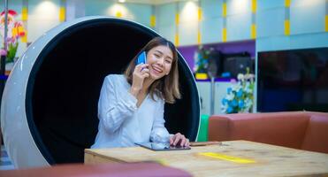 magnifique asiatique affaires femme séance sur moderne rond chaise et en utilisant téléphone à communiquer avec client avec bonheur, numérique commercialisation. photo