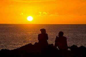 deux gens séance sur le bord de une Roche en train de regarder le le coucher du soleil photo