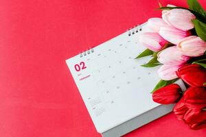 Haut voir, calendrier de février avec brouiller rouge et rose tulipes fleur sur le rouge Contexte bureau, le concept pour la Saint-Valentin journée sur 14e février 2021. fermer, sélectif se concentrer, copie espace pour conception photo