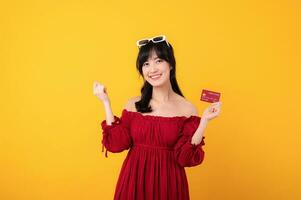 portrait Jeune magnifique asiatique femme content sourire habillé dans rouge vêtements montrant crédit carte isolé sur Jaune Contexte. Payer et achat achats Paiement concept. photo