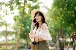 portrait de asiatique Jeune femme voyageur avec tissage chapeau, panier, mobile téléphone et caméra sur vert Publique parc Contexte. périple voyage mode de vie, monde Voyage explorateur ou Asie été tourisme concept. photo