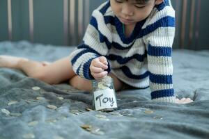 asiatique garçon est assis et met pièces de monnaie dans une verre pot à enregistrer argent à acheter jouets. photo