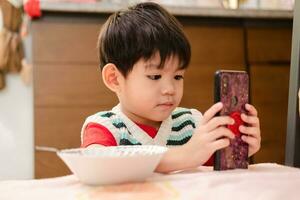 le visage de un asiatique enfant en mangeant tandis que à la recherche à le téléphone photo