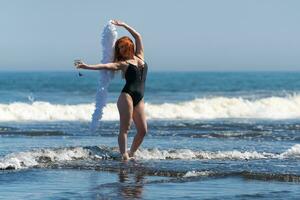 femme dans maillot de bain des stands dans mer vagues, en portant verre de du vin dans tendu main et boa dans autre photo