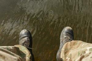 Pour des hommes jambes dans camouflage un pantalon et vieux bottes pendre au dessus le l'eau. photo