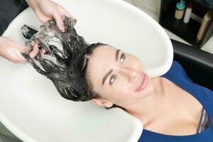 les coiffeurs mains laver longue cheveux de brunette femme avec shampooing dans spécial évier pour shampooing photo