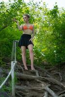 femme permanent dans forêt sur échelle fabriqué de arbre racines. femelle dans Orange bikini Haut et mini jupe photo