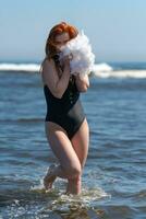 sexy femme dans maillot de bain en marchant dans vagues de océan, en portant boa dans mains et ludique couvrant visage photo