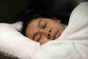 asiatique femme repos couvert avec couverture sur blanc feuilles dans chambre à coucher. femme dormant. paisible femme mensonge dans lit et en gardant yeux fermé tandis que couvert avec couverture. photo