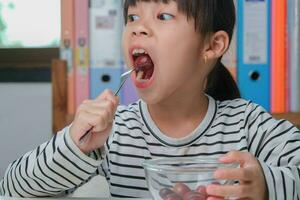 peu fille en mangeant Frais rouge les raisins à Accueil dans le vivant chambre. mignonne Jeune asiatique fille mange en bonne santé des fruits et Lait pour sa repas. en bonne santé nourriture dans enfance photo