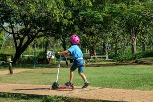 portrait d'une petite fille active faisant du scooter dans la rue dans un parc extérieur le jour d'été. heureuse fille asiatique portant un casque faisant du scooter dans le parc. loisirs actifs et sports de plein air pour enfant. photo