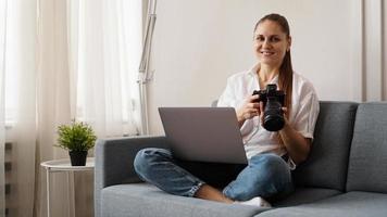 Happy young woman with photo camera using laptop at home