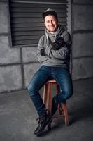 homme avec un appareil photo et une grimace assis sur une chaise dans un studio gris