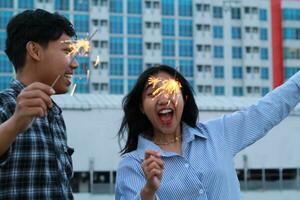 insouciant asiatique Jeune couple en portant cierges magiques célébrer Nouveau année avec ville bâtiment arrière-plan, branché photo