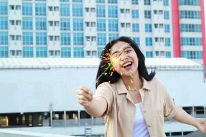 content asiatique Jeune femme en portant cierges magiques célébrer Nouveau année veille dans Extérieur toit Haut avec ville bâtiment Contexte photo