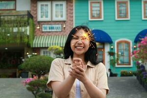Nouveau année veille fête concept. attrayant asiatique Jeune femme en portant cierge magique avec en riant content dans Extérieur jardin de ancien maison Cour photo