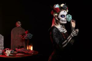 femelle modèle avec sucre crâne faire en haut et bleu des roses habillé dans noir costume de décès comme Père Noël muerte. journée de le mort corps art à célébrer mexicain rituel sur Halloween, culture mythe. photo