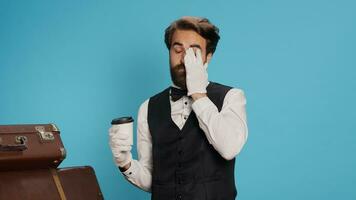 portier pose avec café tasse dans studio, portant blanc gants et sentiment sur de soi travail dans hospitalité secteur. groom employé dans formel tenue en buvant boisson suivant à chariot Sacs pile. photo