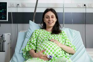 de bonne humeur Enceinte patient attendre pour médecin à début soins de santé examen pendant vérification visite dans hôpital salle. femme avec ventre grossesse mensonge dans lit, souriant à caméra avant chirurgie photo