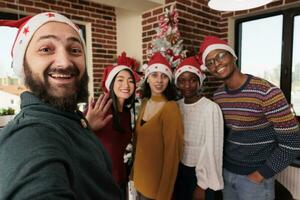 de bonne humeur des employés équipe portant Père Noël Chapeaux prise selfie tandis que travail dans décoré entreprise bureau. collègues de travail posant pour téléphone intelligent photo tandis que célébrer Noël veille dans cotravail espace