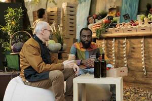 Masculin client en buvant fait maison du vin avec local vendeur et séance ensemble à Les agriculteurs marché tableau. Jeune homme client profiter nourriture et du vin dégustation, visite biologique l'automne festival. photo