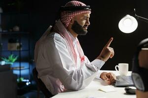 vue de côté coup de islamique gars portant traditionnel Vêtements assise à le sien bureau ayant une conférence appel sur le sien portable. image spectacles arabe pigiste absorbé dans une conversation sur le sien personnel ordinateur. photo