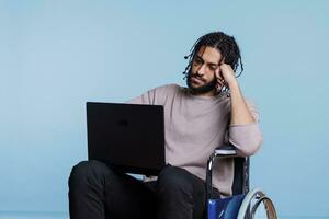 fatigué arabe homme dans fauteuil roulant dactylographie sur portable tandis que travail à distance. Jeune épuisé pigiste Logiciel développeur avec physique mettre en œuvre en cours d'analyse code sur portable ordinateur photo