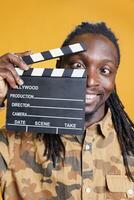 Jeune adulte en portant filmographie ardoise, posant sur de soi dans de face de caméra dans studio plus de Jaune Contexte. africain américain homme travail dans cinématographie production industrie photo