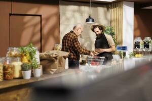 personnes âgées homme à check-out dans zéro déchets boutique, achat localement grandi respectueux de la nature des légumes de amical magasinier. plus âgée client achat biologique produits chimiques gratuit les courses photo