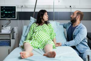 femme avec grossesse mensonge dans hôpital quartier lit avec mari à côté de sa attendre pour enfant livraison. patient en train de préparer pour la main d'oeuvre ayant douleur contractions, demander pour médecin à viens et début chirurgie photo