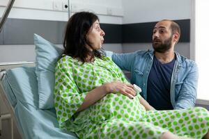 Enceinte femme Aller dans la main d'oeuvre dans hôpital salle, mari permanent à côté de sa réconfortant. patient avec grossesse en portant mains sur ventre Faire respiration exercices tandis que ayant douloureux contractions photo