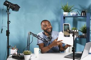 africain américain en ligne blogueur vivre diffusion révéler concours en utilisant téléphone intelligent sur trépied. souriant vlogger Parlant et en portant cadeau boîte tandis que enregistrement vidéo sur mobile téléphone photo