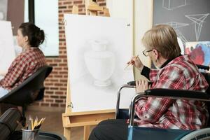 Sénior femme fauteuil roulant utilisateur dessin avec crayon sur toile, assister art Des classes pour gens avec invalidité, réduire stress dans Créatif espace. Créatif thérapie pour désactivée adultes photo