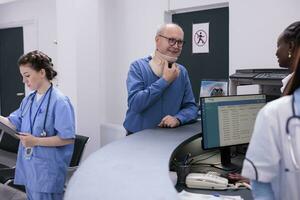 blessé Sénior patient avec cervical cou collier permanent à hôpital accueil compteur discuter à propos rendez-vous avec réceptionniste. la personne avec médical entretoise attendre à rencontrer avec traumatologue. photo