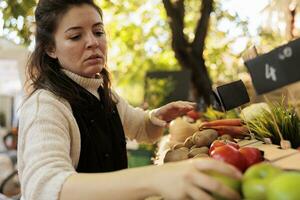 vendeur organiser Frais biologique produire à Les agriculteurs marché rester, femme vendeur avec tablier. Frais biologique des fruits et légumes sur supporter à Extérieur Les agriculteurs marché. femelle agriculteur vente fait maison produire. photo