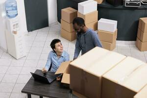 Haut vue de diverse équipe discuter à propos livraison logistique tandis que à la recherche à cargaison Stock sur portable ordinateur, ouvriers en train de préparer clients paquets. des employés mettre ordres dans carton des boites dans entrepôt photo